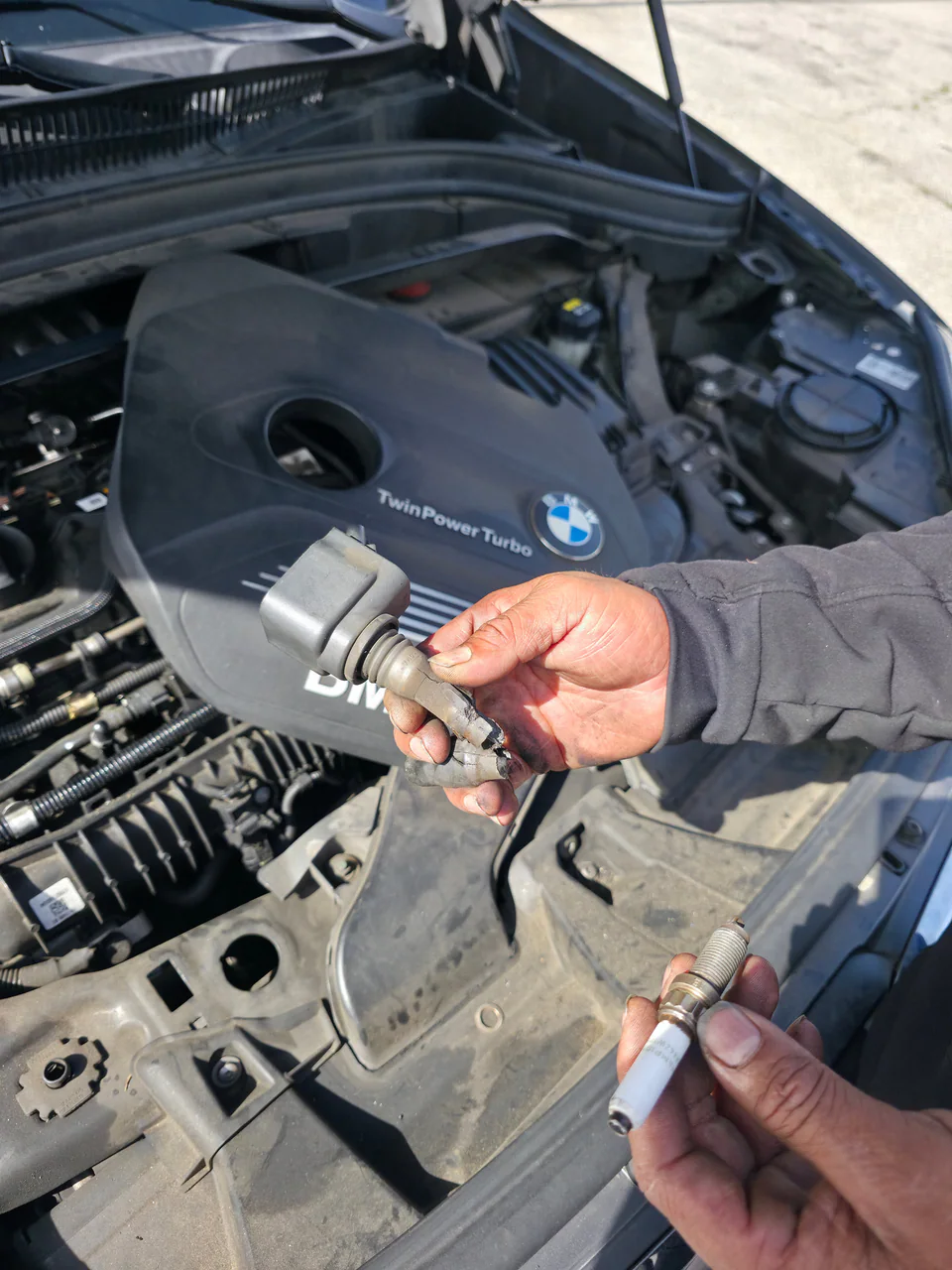 Hands holding an ignition coil and spark plug above a BMW engine at the shop